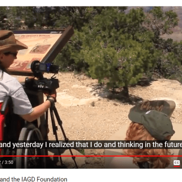 A female faulty member with brown hair and a cowboy hat, adjust's her camera equipment from her wheelchair and chats with a student sitting on the ground next to her on the rim of the Grand Canyon.