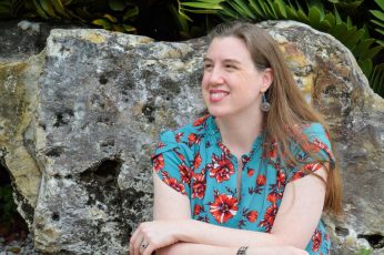 Anita Marshall sits in front of a gray rock and tropical foliage smiling off the the side of the camera. She is a fair skinned woman with long dark blonde hair.