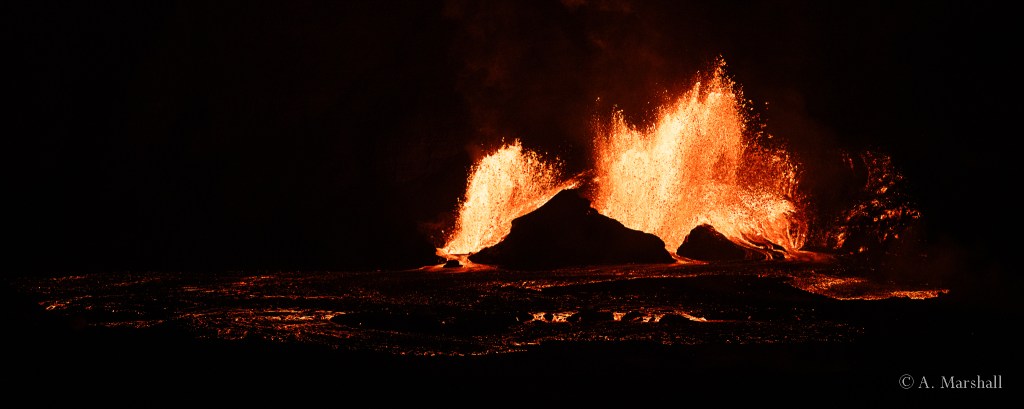 Fire fountains of lava erupt from a small crater at night.