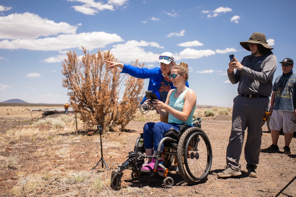 Anita Marshall wears a blue field shirt and points away toward the distance while looking over the shoulder of a student in a wheelchair operating a drone.