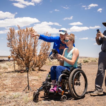 Anita looks over the shoulder and points away in the distance as a student flies a drone to an inaccessible outcrop.